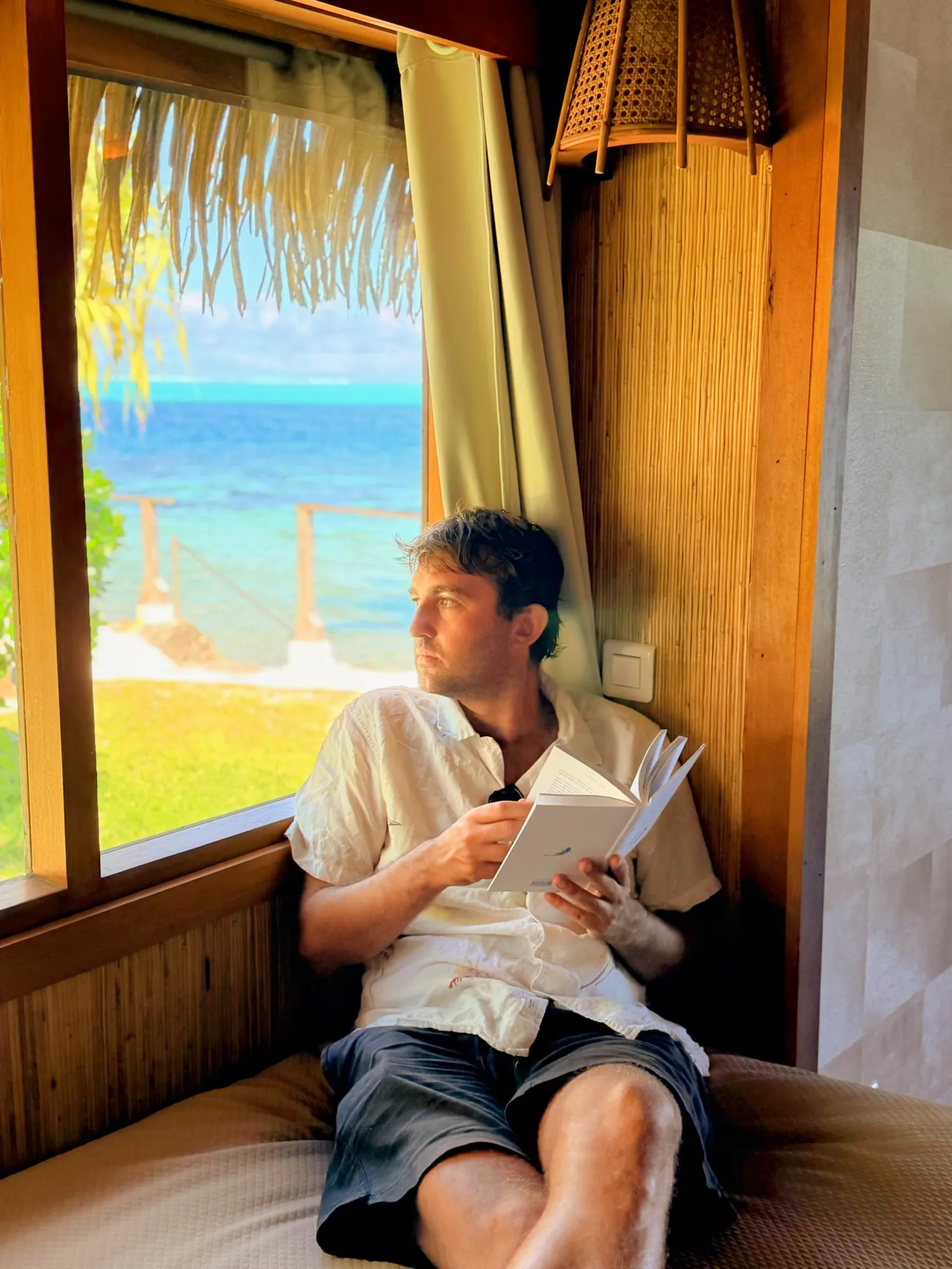 Homme lisant près de la fenêtre avec vue sur le lagon dans un bungalow, Hôtel Le Mahana, Huahine